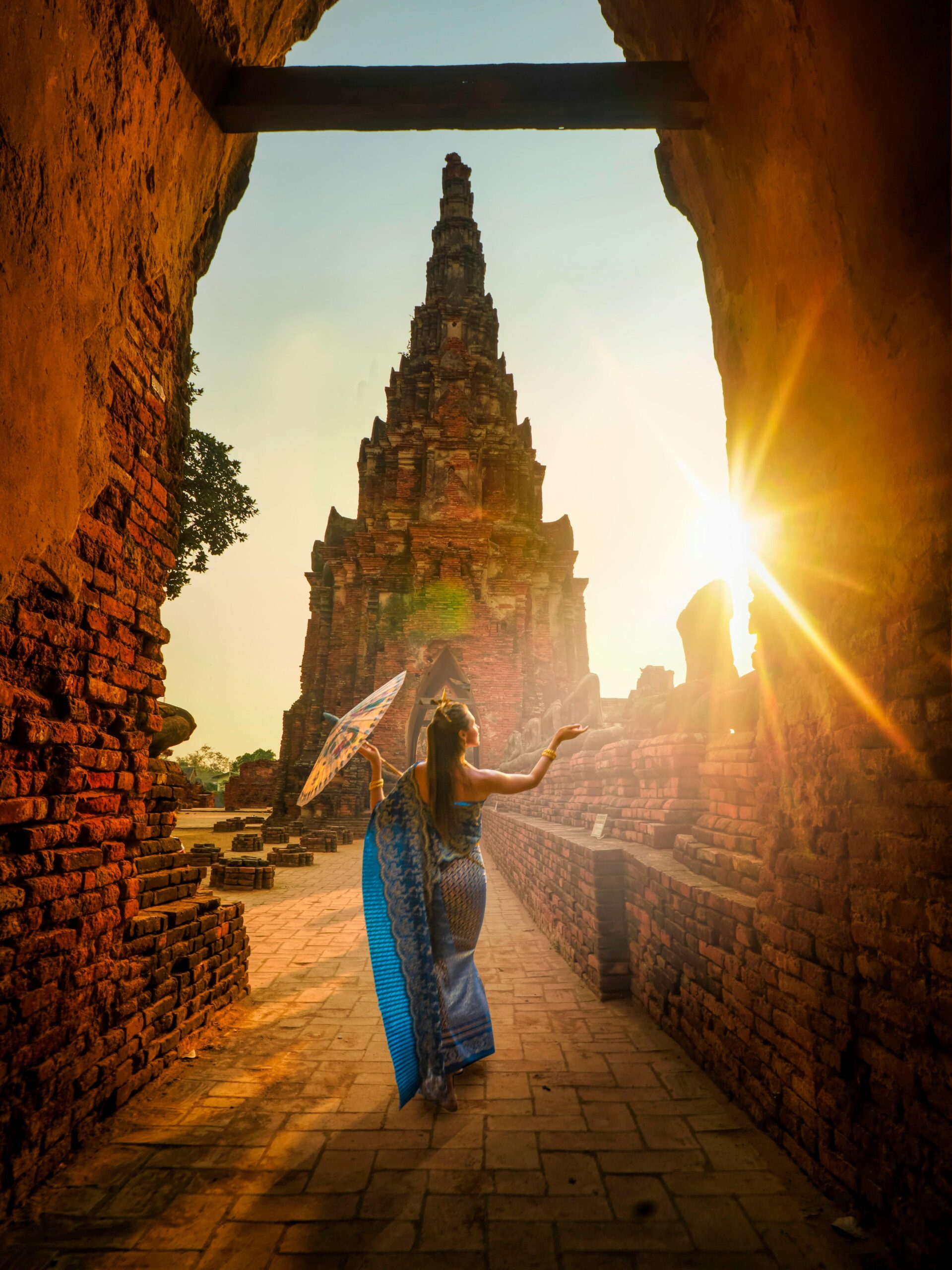 A woman in traditional wear stands with an umbrella at a historic Ayutthaya temple during sunset.